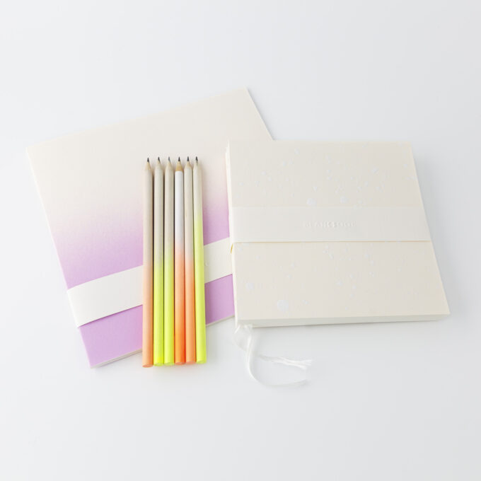 Liggande skrivbok med pennor i övertoningar av lysande färger och anteckningsbok i taktilt mönster med bokmärkesband.